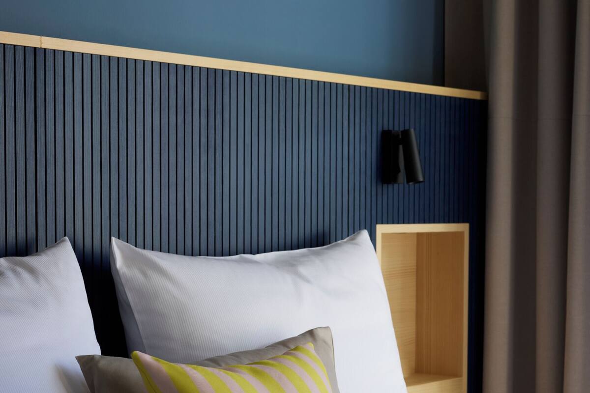 A hotel room with a bed, pillows, a headboard, a bedside table, and a lamp.