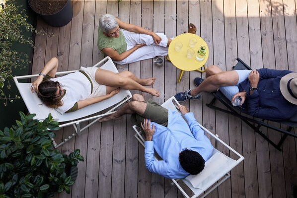 Una terraza de madera con cuatro sillas reclinables, una mesita redonda con bebidas y plantas en macetas.