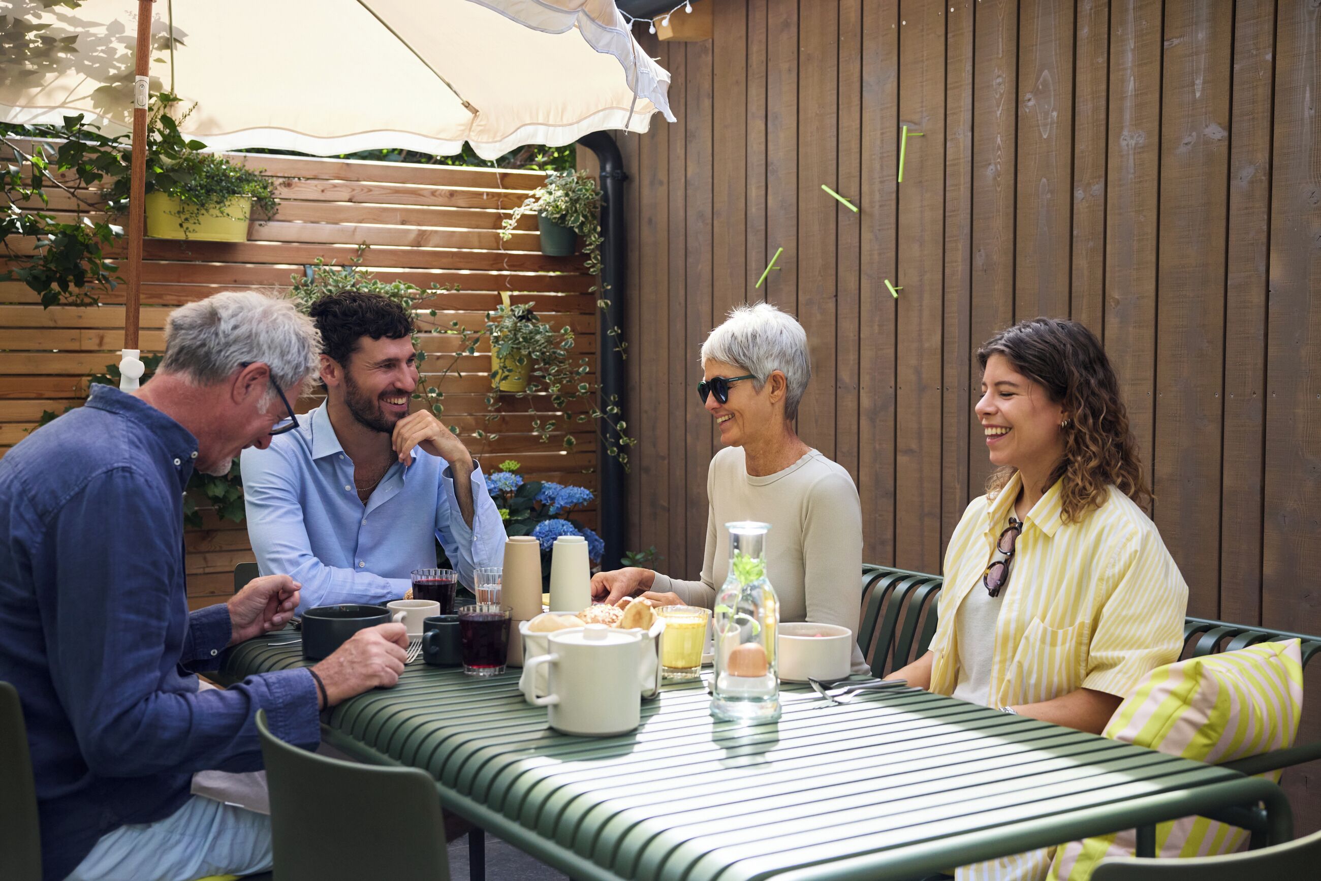 Um grupo de pessoas sentadas em uma mesa ao ar livre, com um cercado de madeira e vasos de plantas ao fundo.