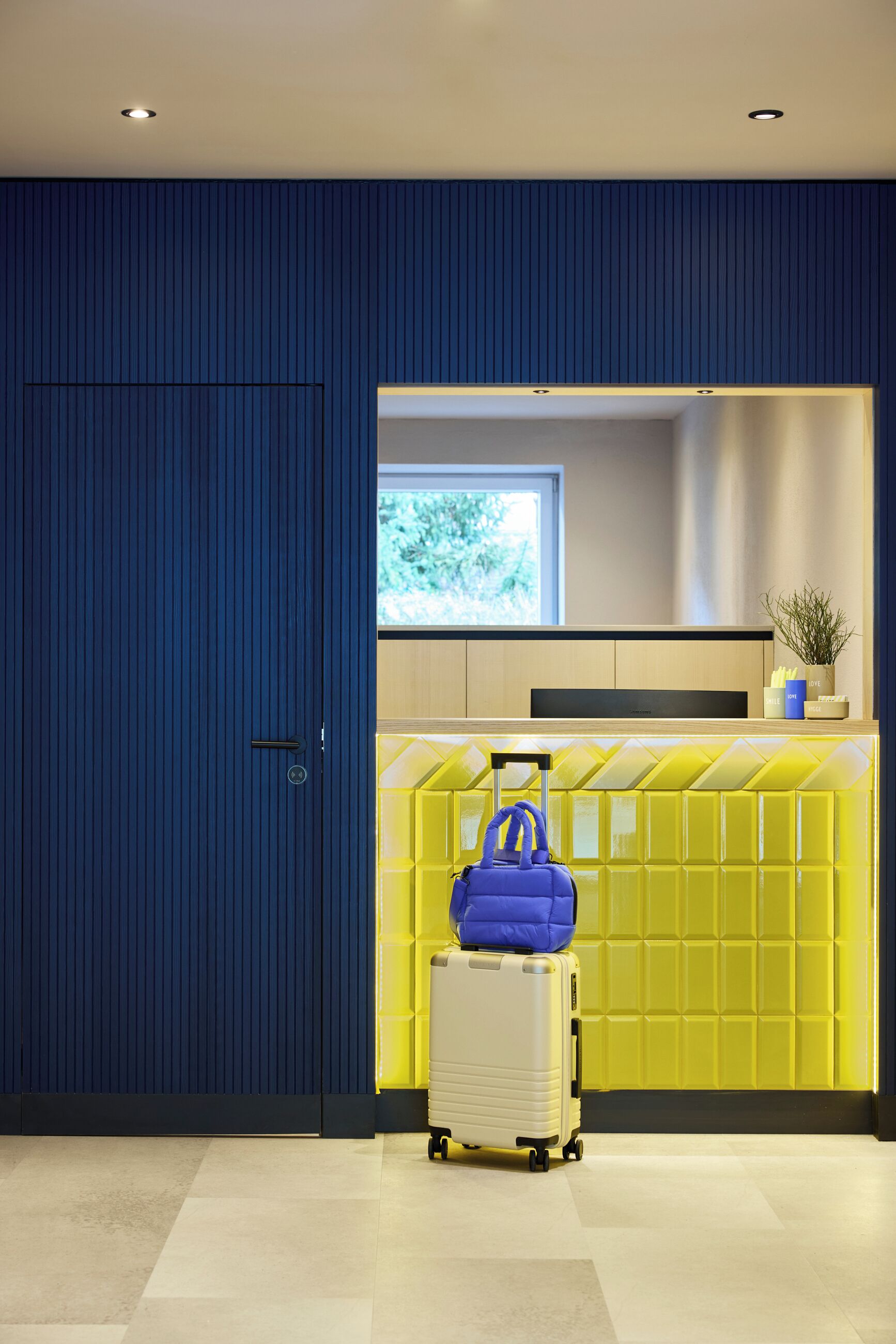 Una habitación con una pared azul, una pared amarilla y un ventanal. Hay una maleta y un bolso azul en el suelo.