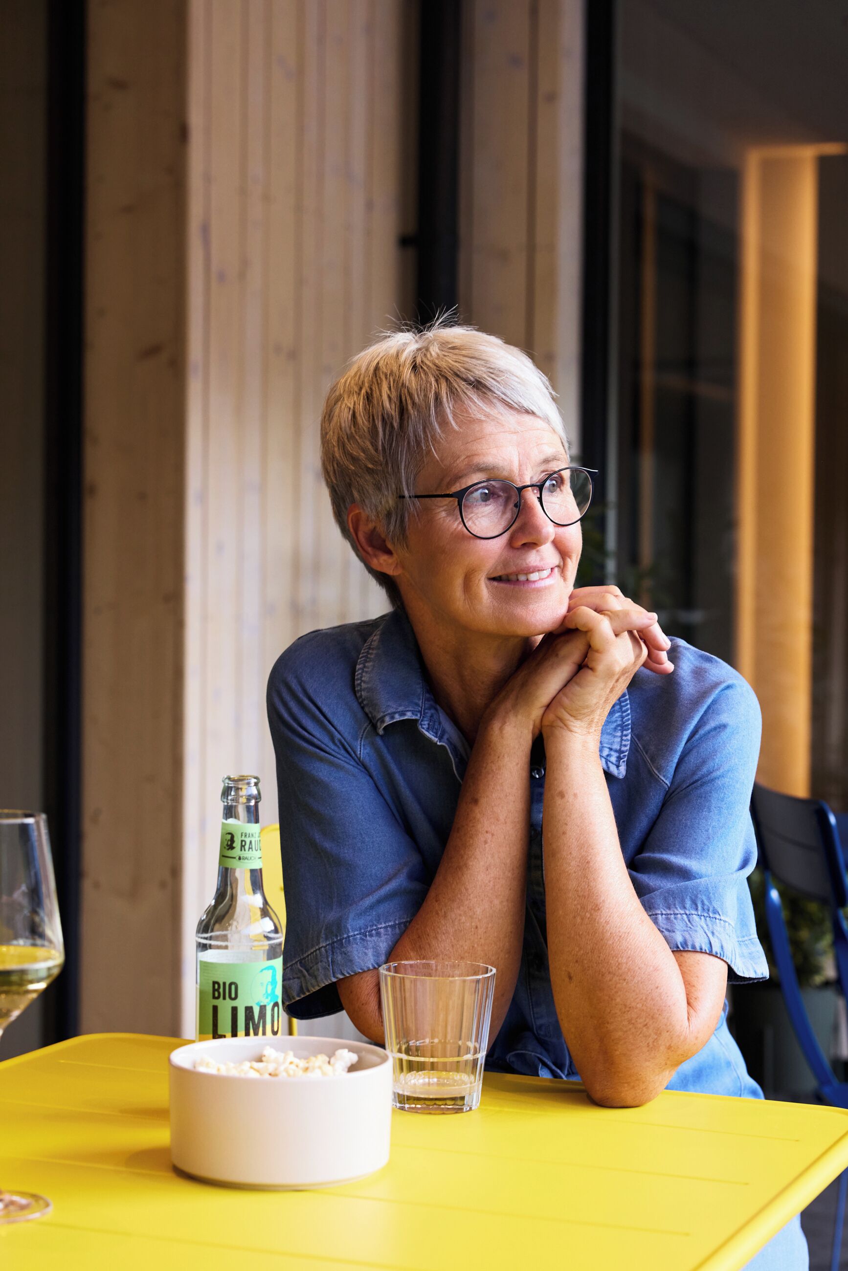 A person seated at a yellow table with a bottle of Bio Limo, a bowl of popcorn, and a glass.