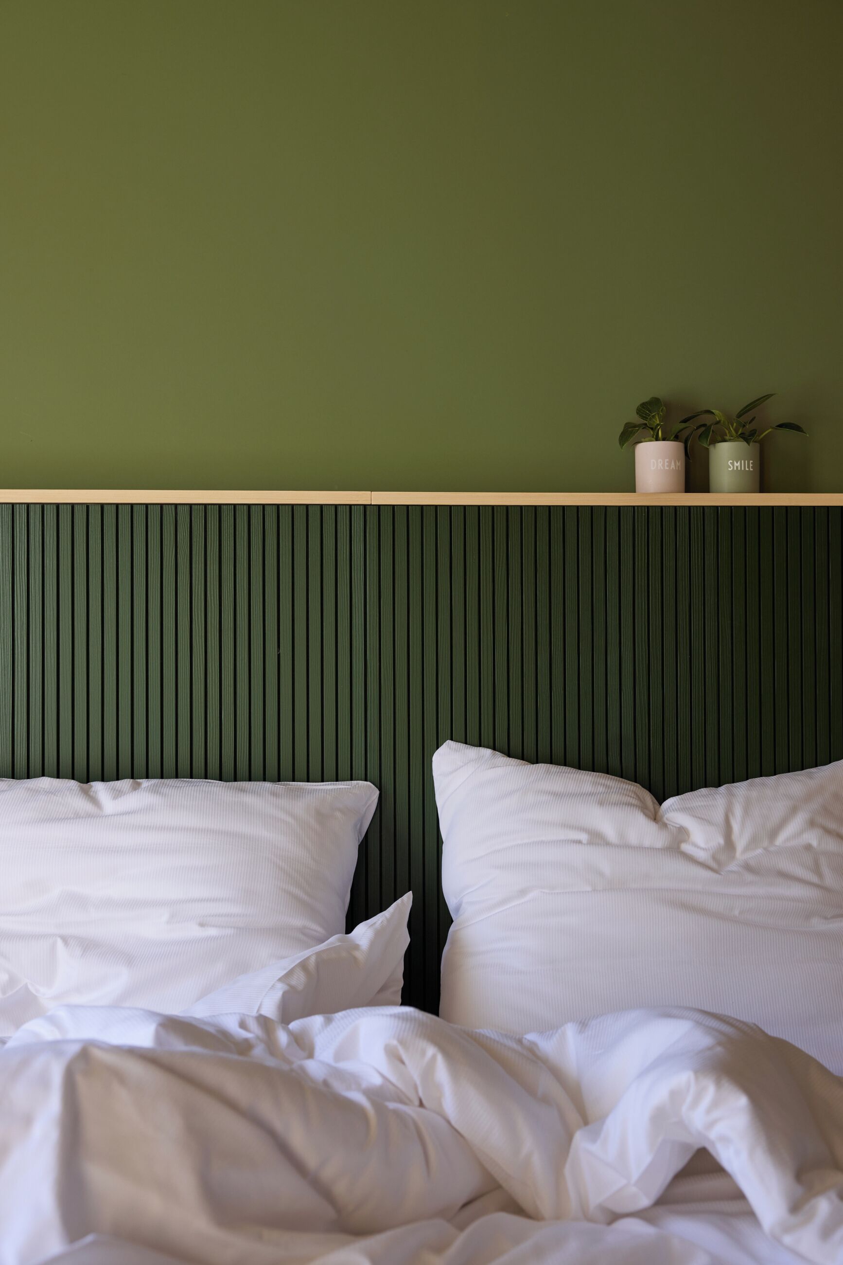 A bed with white bedding and pillows, a green wall, and two potted plants on a shelf above the bed.