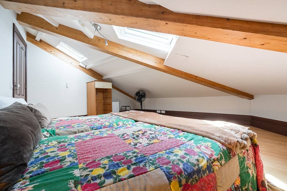 A bedroom with a sloped ceiling, wooden beams, a bed with a colorful quilt, a bedside table, and a fan.