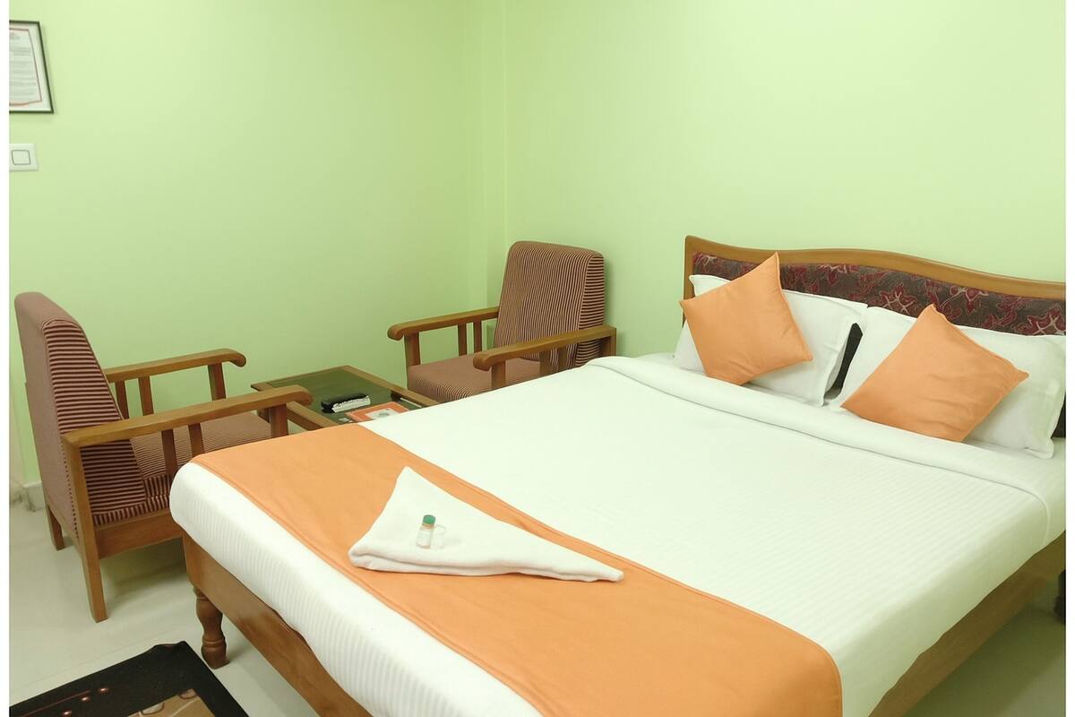 A neatly made bed with an orange blanket and pillows, a wooden chair, a small table, and a framed certificate on the wall.