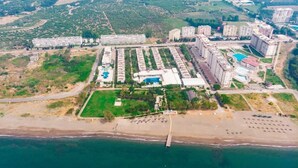 An aerial view of a coastal resort with multiple high-rise buildings, a central pool area, and a beach with sun loungers. - Doramar Resort Aqua Tatilkoyu (Erdemli)