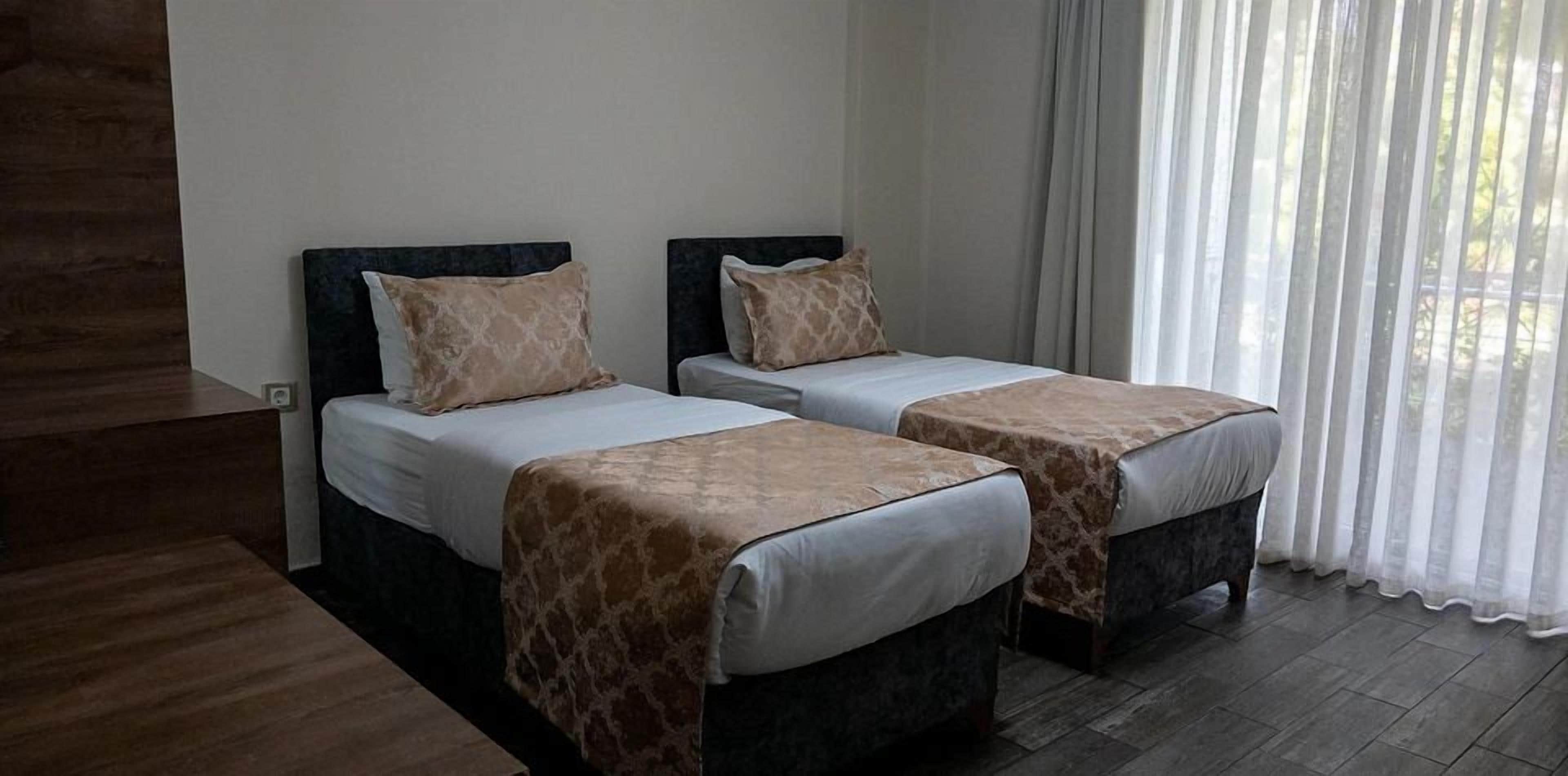 A hotel room with two beds, a wooden floor, and a view through sheer curtains.