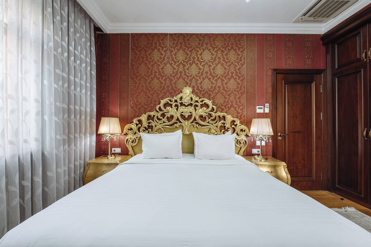 Un grand lit avec une tête de lit dorée, deux lampes de chevet et un mur orné d’un motif rouge.