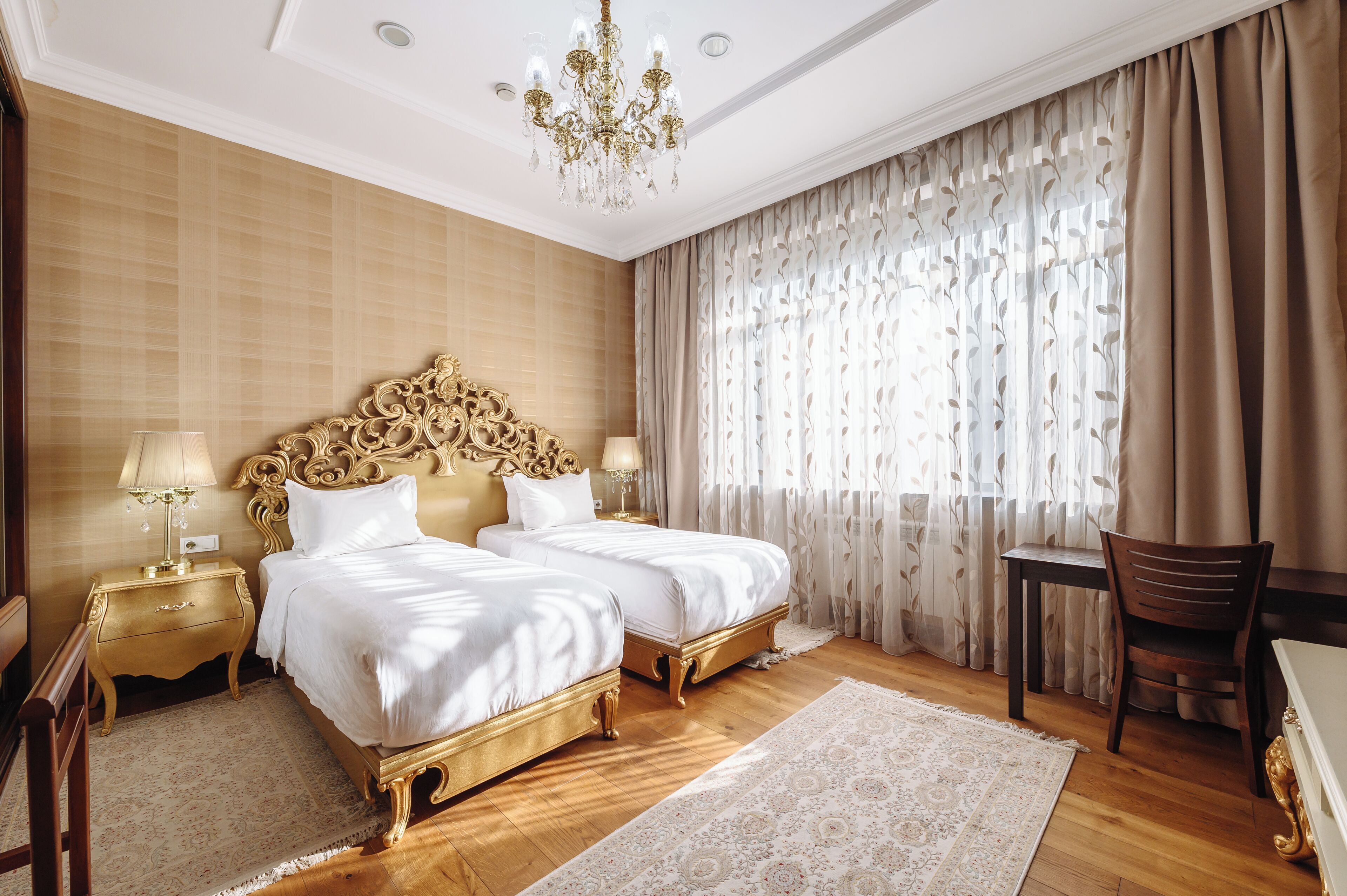 Habitación de hotel con dos camas, un candelabro, suelo de madera y un ventanal con cortinas.