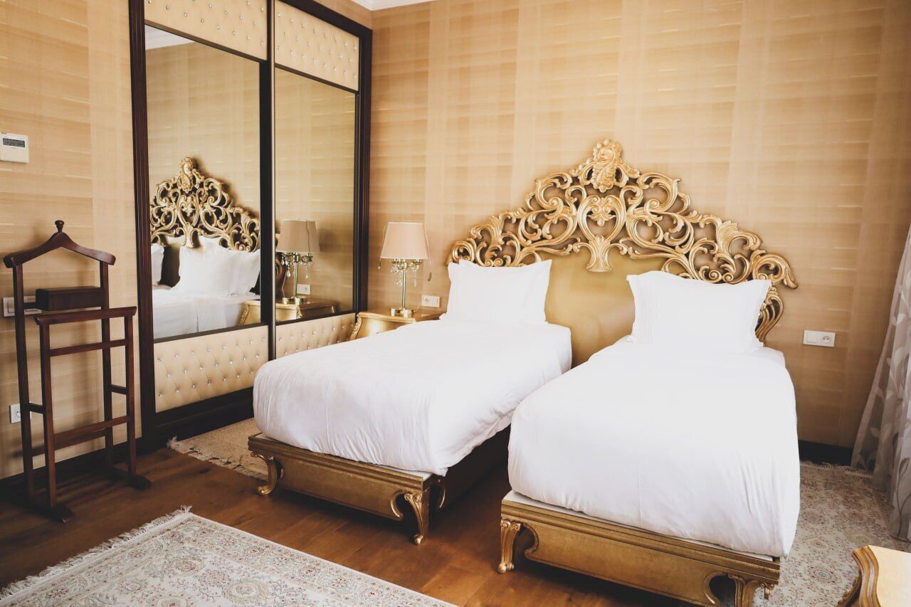 A double bed with a gold headboard and white bedding in a room with wooden flooring and a large mirror.