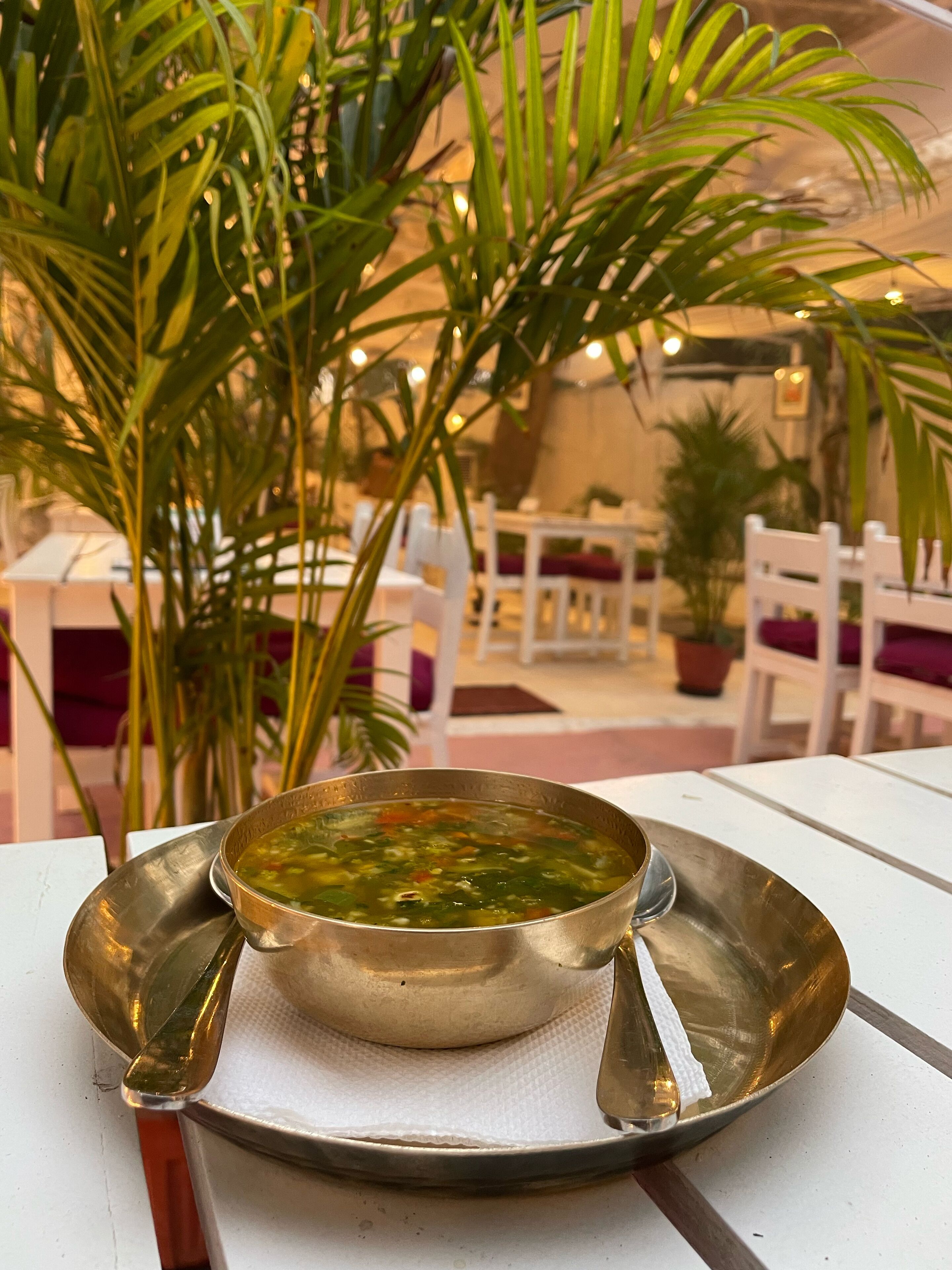 A table with a metal bowl of soup, white chairs, and potted plants.