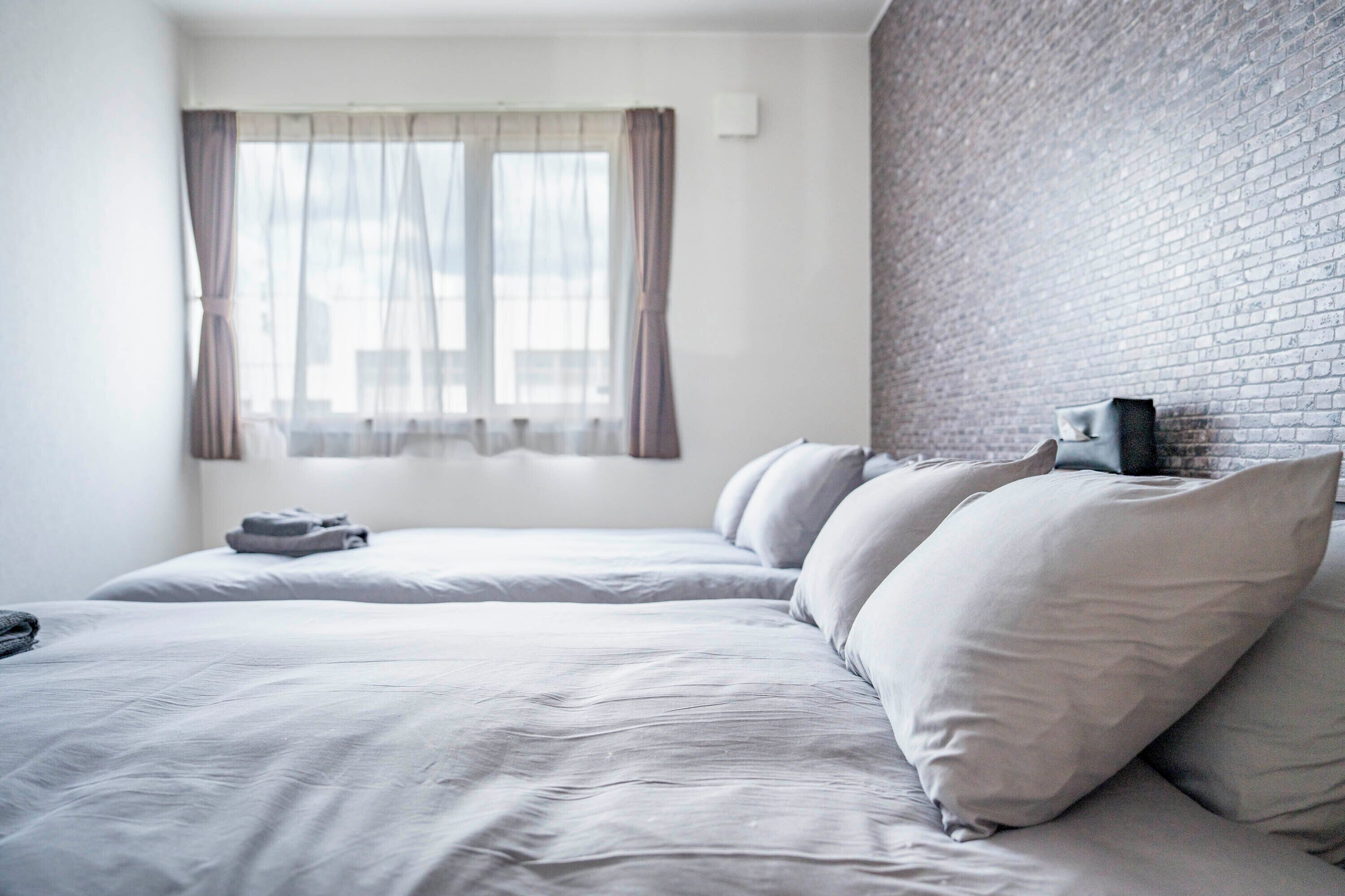 A hotel room with two beds, a brick wall, and a window with sheer curtains.