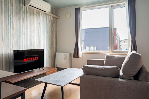 A modern living room with a flat-screen TV displaying 'NETFLIX', a sofa, a coffee table, and a window with curtains. - LAMBENT (Kamifurano)