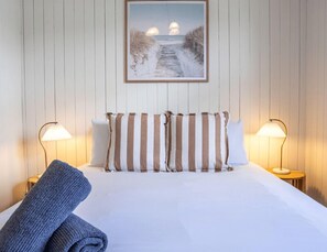 En pænt redt seng med stribede puder, to sengeborde med lamper, og et billede af en strandscene på væggen.