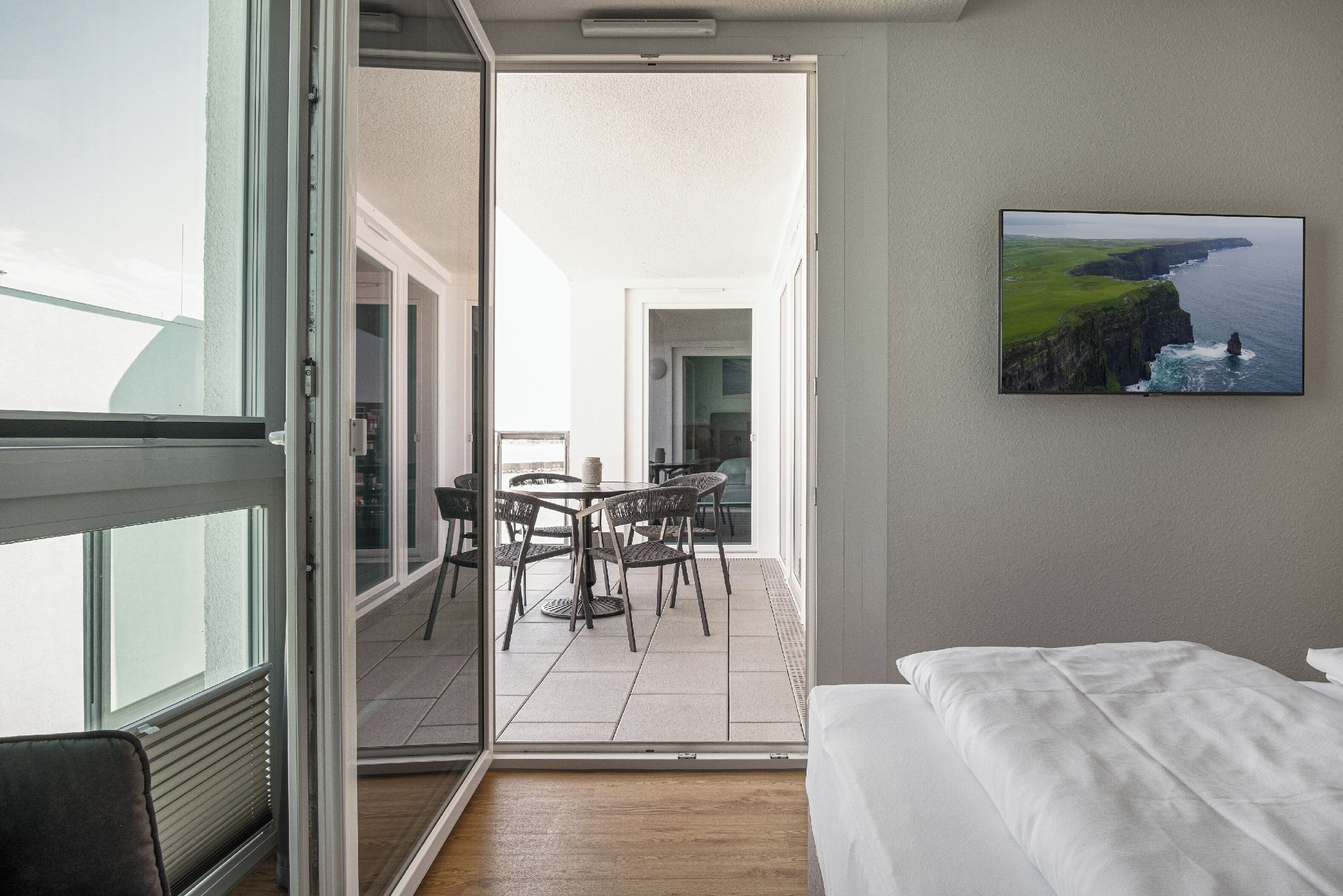 Een hotelkamer met een bed, uitzicht op een balkon en een schilderij van een kustlandschap.