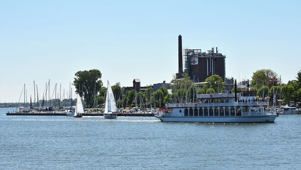 Ein Fluss mit einem großen weißen Boot, das am Steg liegt, umgeben von Segelbooten und einem Gebäude mit Schornstein im Hintergrund.