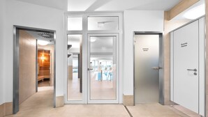 A modern hotel lobby with a glass door leading to a pool area, a reception desk, and a sign indicating "Umkleide Damen" (women's changing room).
