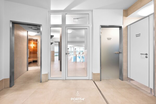 Een moderne hotellobby met een glazen deur naar een zwembadgedeelte, een receptiebalie en een bordje met de tekst 'Umkleide Damen'.