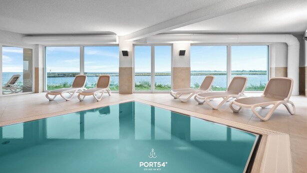 A modern indoor swimming pool with clear views of the water outside, surrounded by lounge chairs.