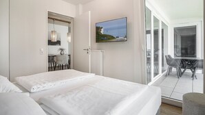 A hotel room with two beds, a dining area with a table and chairs, a large window with a view of the sea, and a painting on the wall.