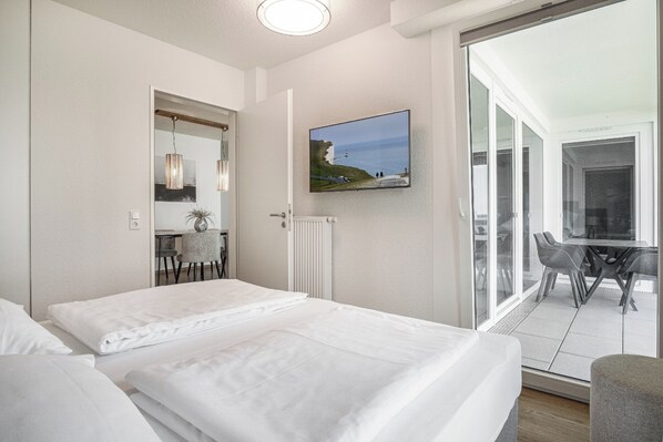 Quarto de hotel com duas camas, área de jantar com mesa e cadeiras, uma janela grande com vista para o mar e um quadro na parede.