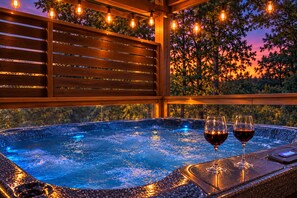 A hot tub with two glasses of red wine, surrounded by wooden slats and string lights.