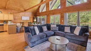 A cozy cabin interior with a large wooden ceiling, a sectional sofa, a dining area, and large windows offering a view of the outdoors.