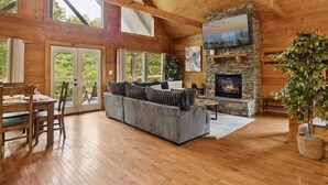 A spacious living room with a stone fireplace, wooden beams, and large windows.