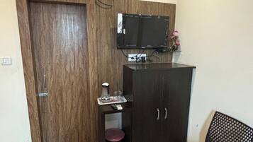 Una habitación pequeña y bien iluminada, con una puerta de madera, un televisor montado en la pared, un mueble con una mesita, un cubo de basura y una silla.