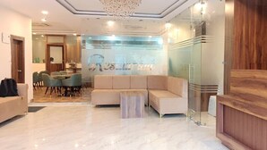 Zona de recepción de hotel con un mostrador de madera, un sofá color beige y un separador de vidrio con el nombre del hotel.