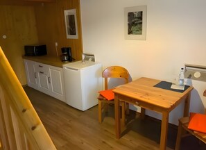 A compact kitchen with a refrigerator, coffee maker, and a microwave. There is a wooden dining table with two chairs, one with an orange cushion.
