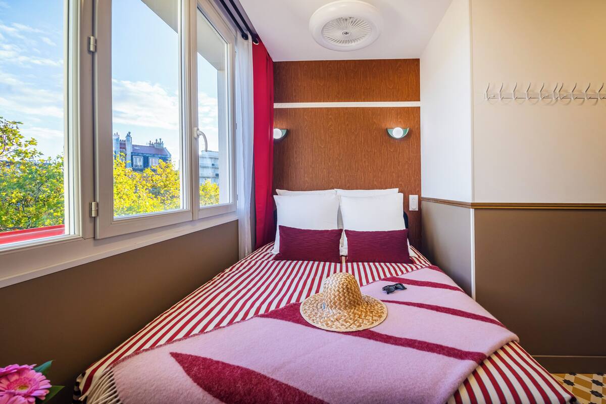 A hotel room with a large window, a bed with striped bedding, a hat on the bed, and a wall-mounted light fixture.