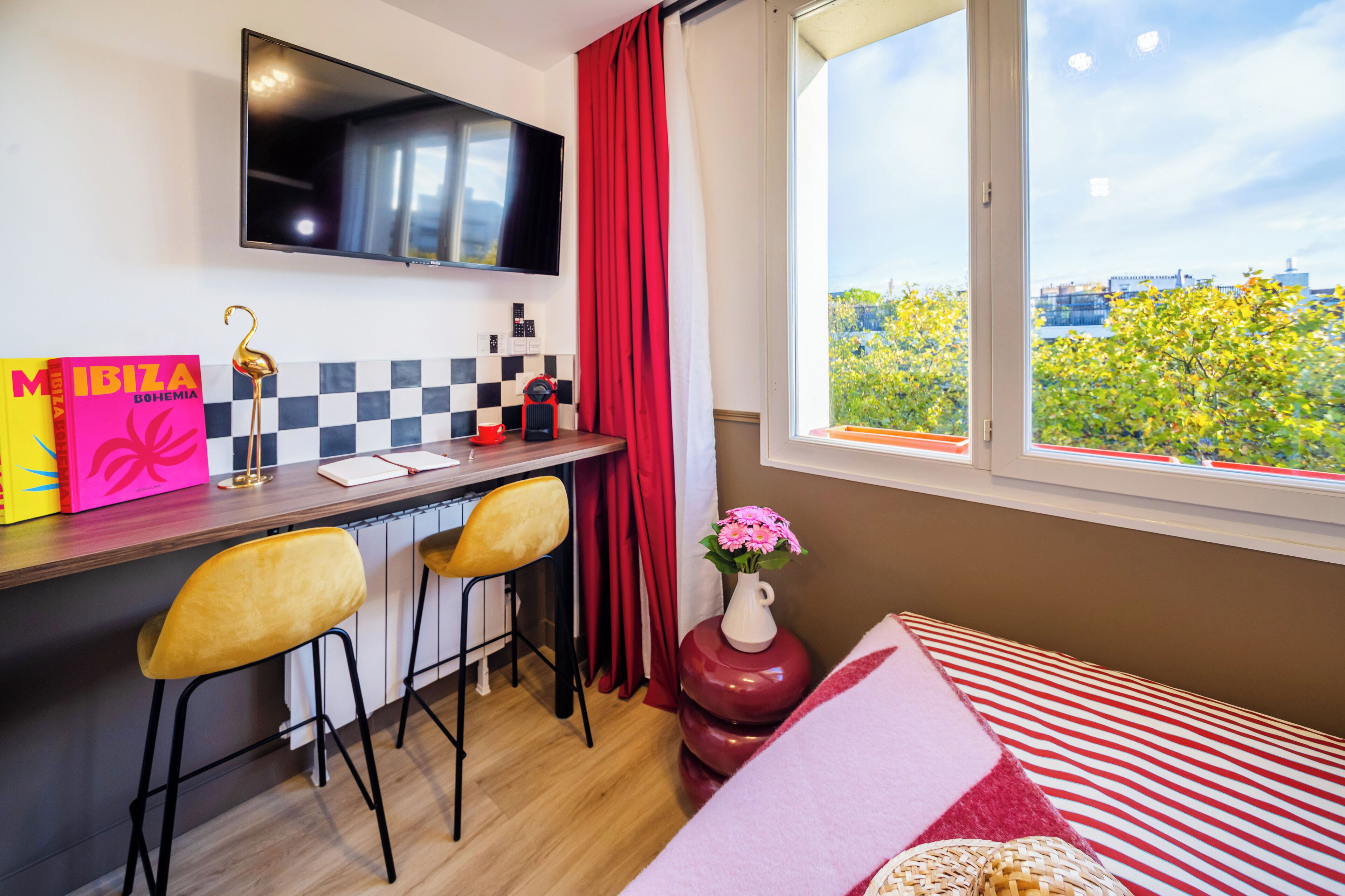 A modern room with a desk, two chairs, a TV, a window with a city view, and a bed with a striped blanket.