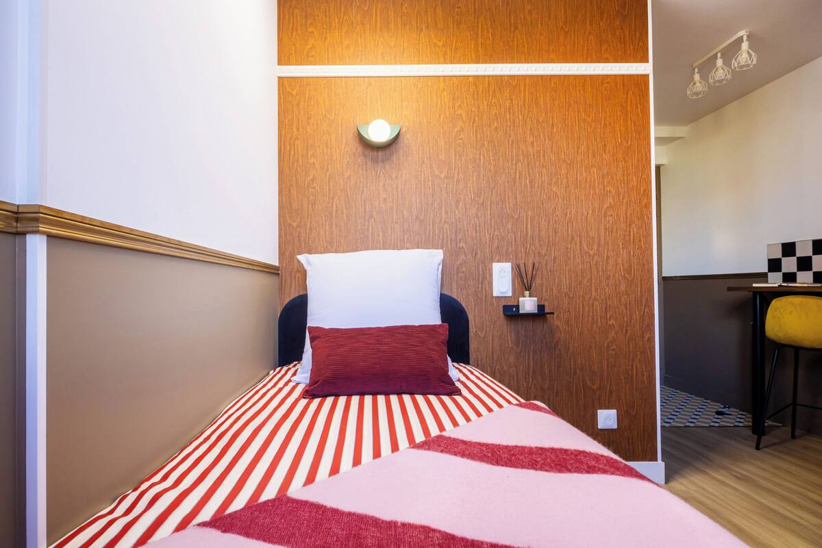 A hotel room with a bed, a wooden accent wall, a small table with a chair, and a wall-mounted light fixture.