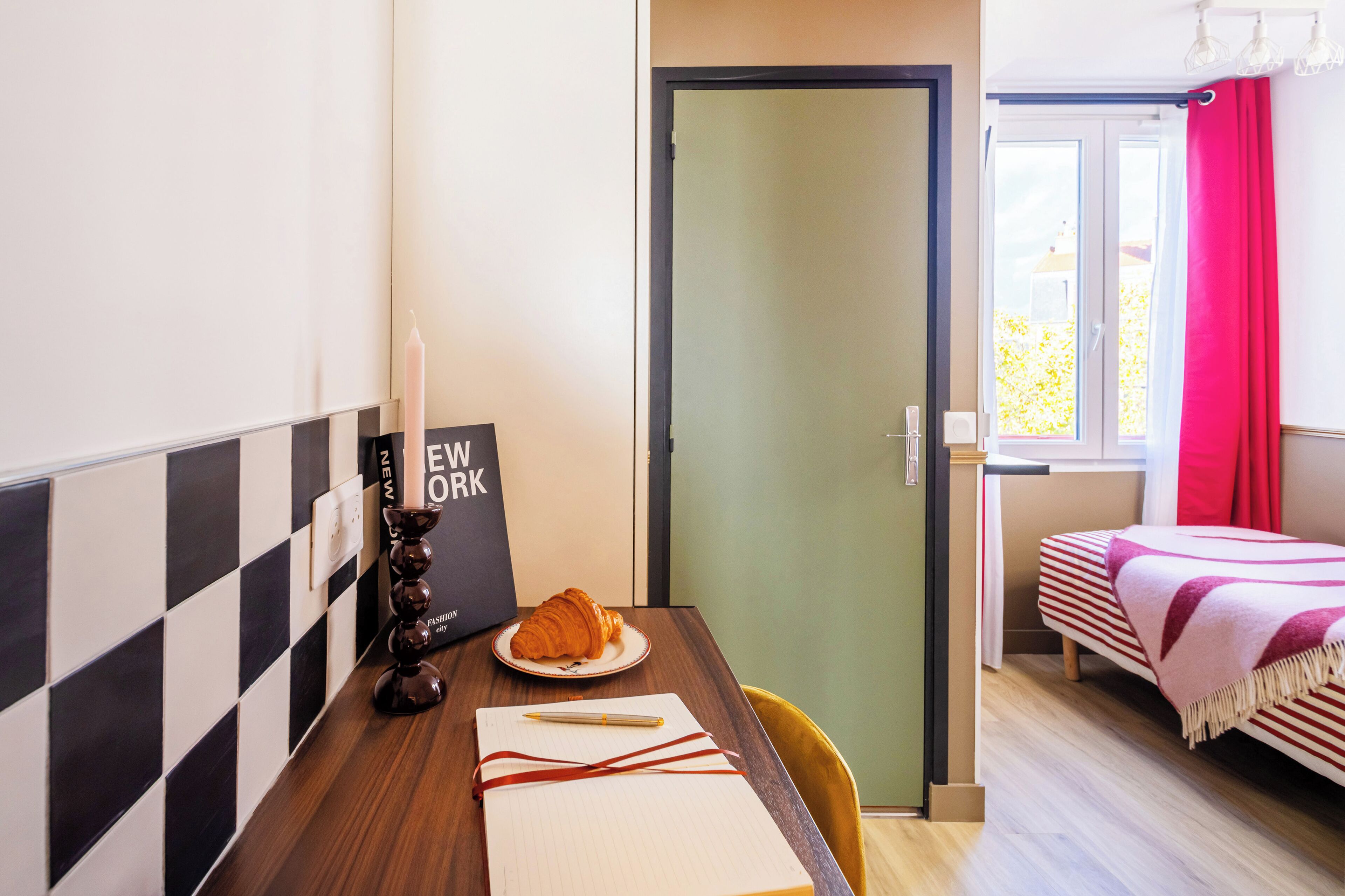 Una habitación pequeña con una mesa de madera, una cama con ropa de cama a rayas, una puerta y una ventana con cortinas.