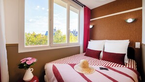 Une chambre d’hôtel avec une grande fenêtre, un lit avec une literie à rayures, un vase rempli de fleurs et un chapeau posé sur le lit.