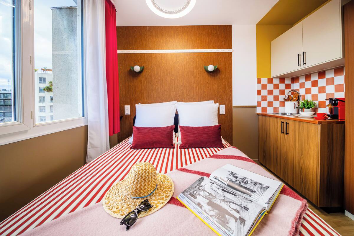 A hotel room with a bed, a small kitchenette, a window, and a view of buildings outside.