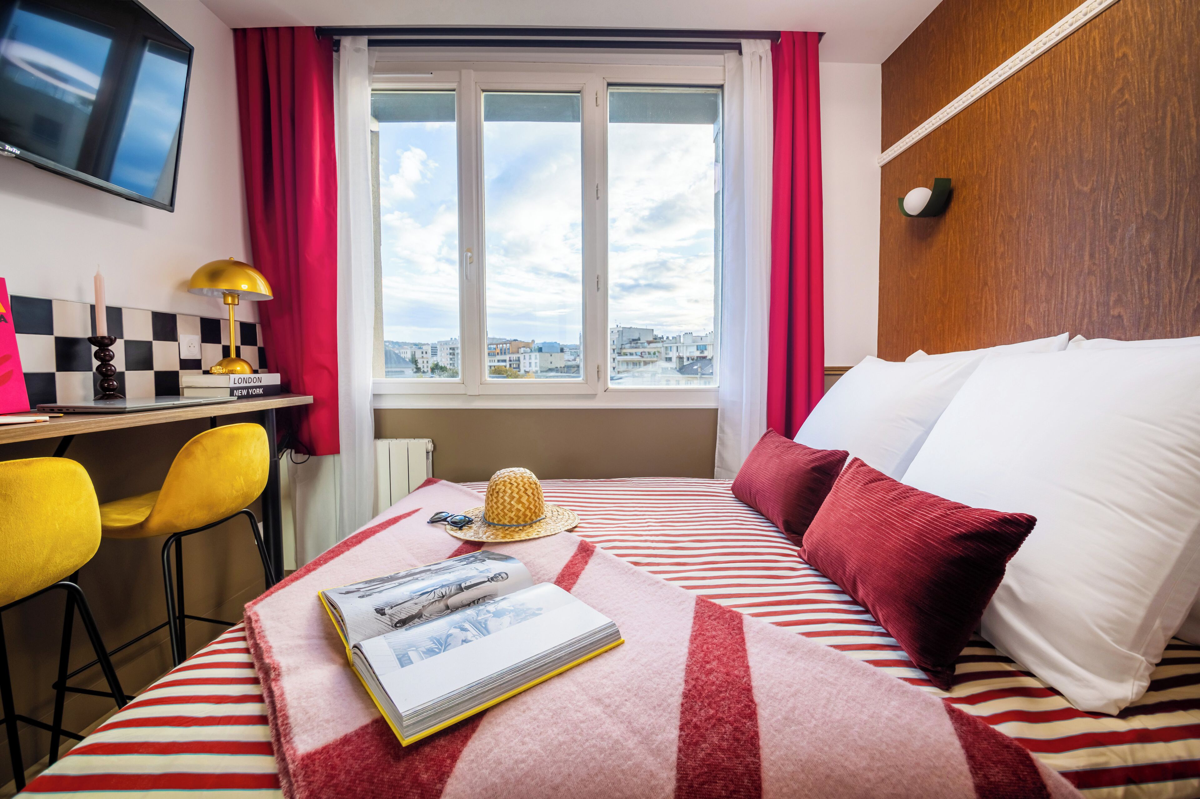 A hotel room with a bed, a desk with a lamp, a chair, a window with a view, and a television on the wall.