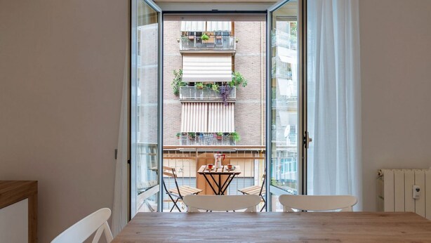A dining area with a wooden table, white chairs, and a view of a balcony with plants and a table. - GuestHost - Trastevere Apartment Roma Portuense (Rome)