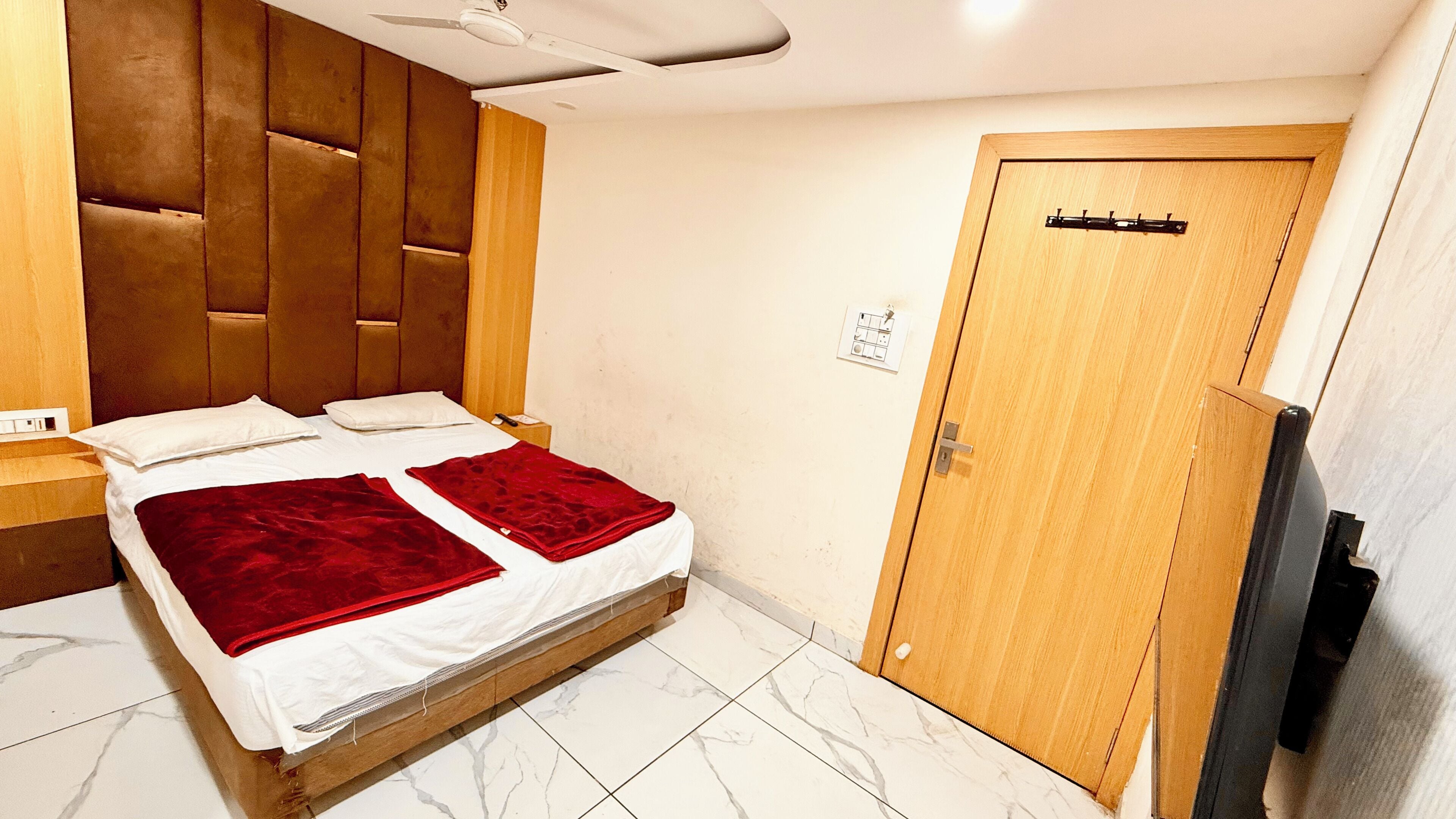 A hotel room with a wooden headboard, a bed with two red blankets, a wooden door, and a television on the wall.