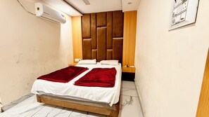 Habitación de hotel pequeña y estrecha con cabecera de madera, cama con sábanas blancas y mantas rojas, mesita de noche de madera y aire acondicionado instalado en la pared.