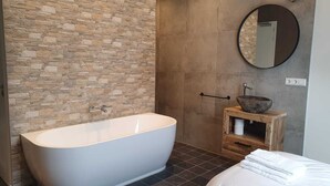 Un baño moderno con una bañera independiente, una pared de piedra, un tocador de madera con lavabo y un espejo redondo.