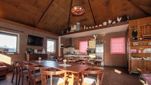 A spacious dining area with wooden tables and chairs, a kitchen with red and white checkered curtains, and a wooden ceiling.