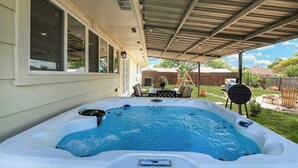 A covered outdoor hot tub area with a patio table and chairs, a barbecue grill, and a wooden fence in the background.