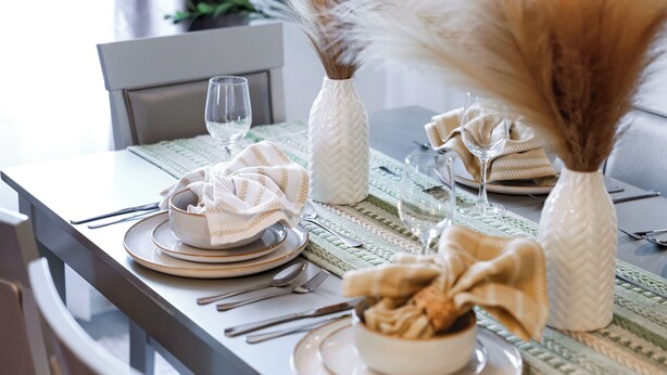 Een eettafel gedekt met borden, kommen, bestek en decoratieve elementen zoals een vaas en pluisgras.