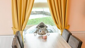 A dining table with chairs, yellow curtains, a 'WELCOME' sign, a candle, and a view of a garden.