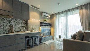 A modern kitchen with grey cabinets, a tiled backsplash, and a central island with stools. There is a sofa and a small side table with a plant in the living area. - Steps to Kamala Beach Rooftop Pool  C138 (Kamala)