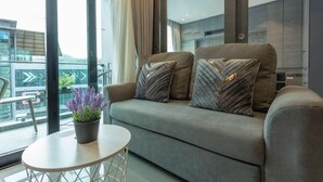 A modern living room with a grey sofa, two patterned cushions, a small round table with a potted plant, and a view of a cityscape through large windows. - Steps to Kamala Beach Rooftop Pool  C138 (Kamala)