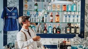 Een bar met flessen drank op de planken, een Chelsea voetbalshirt en een persoon die bedient.