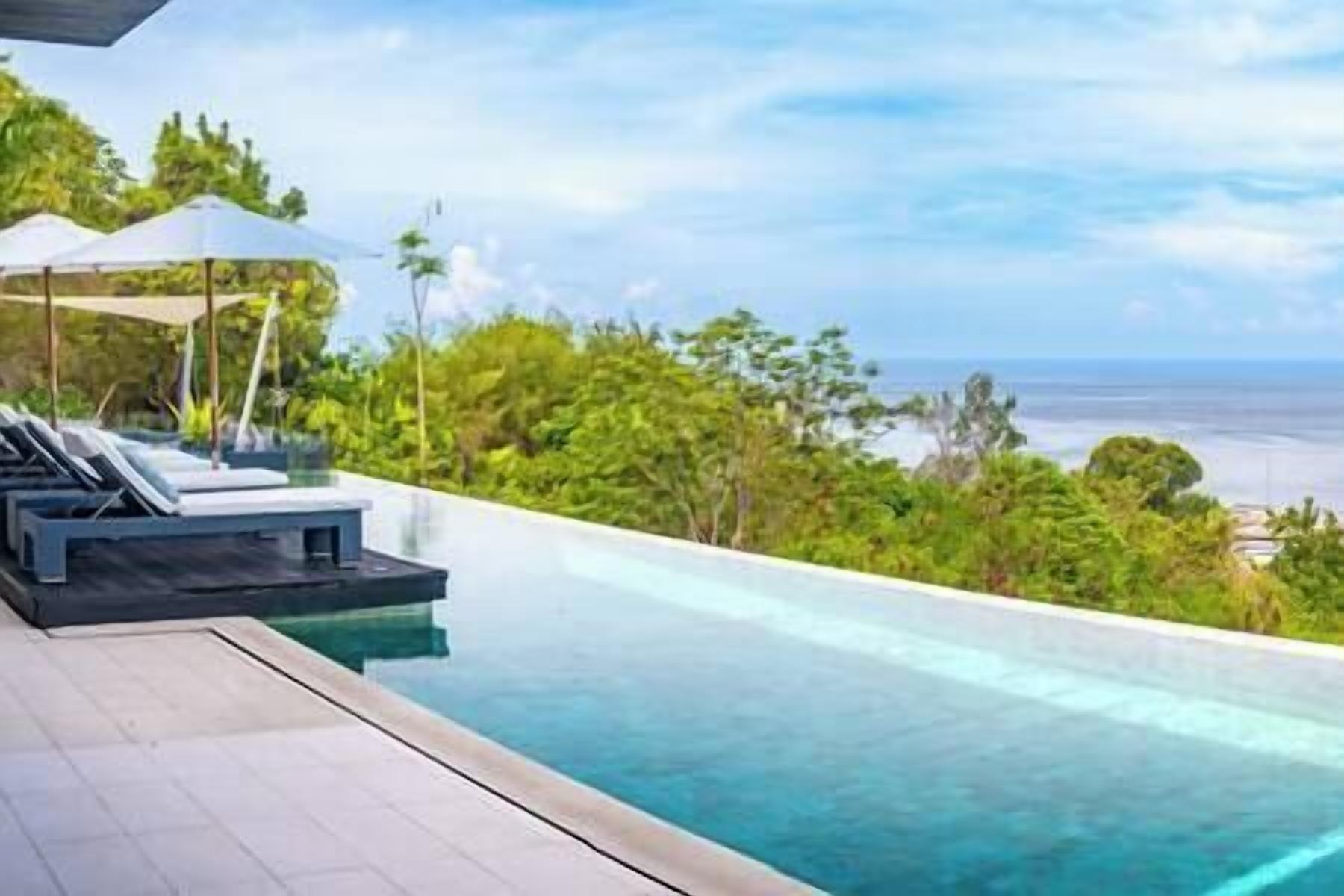 Piscina infinita moderna con sillones de descanso y sombrillas, con vista a una playa y exuberante vegetación.