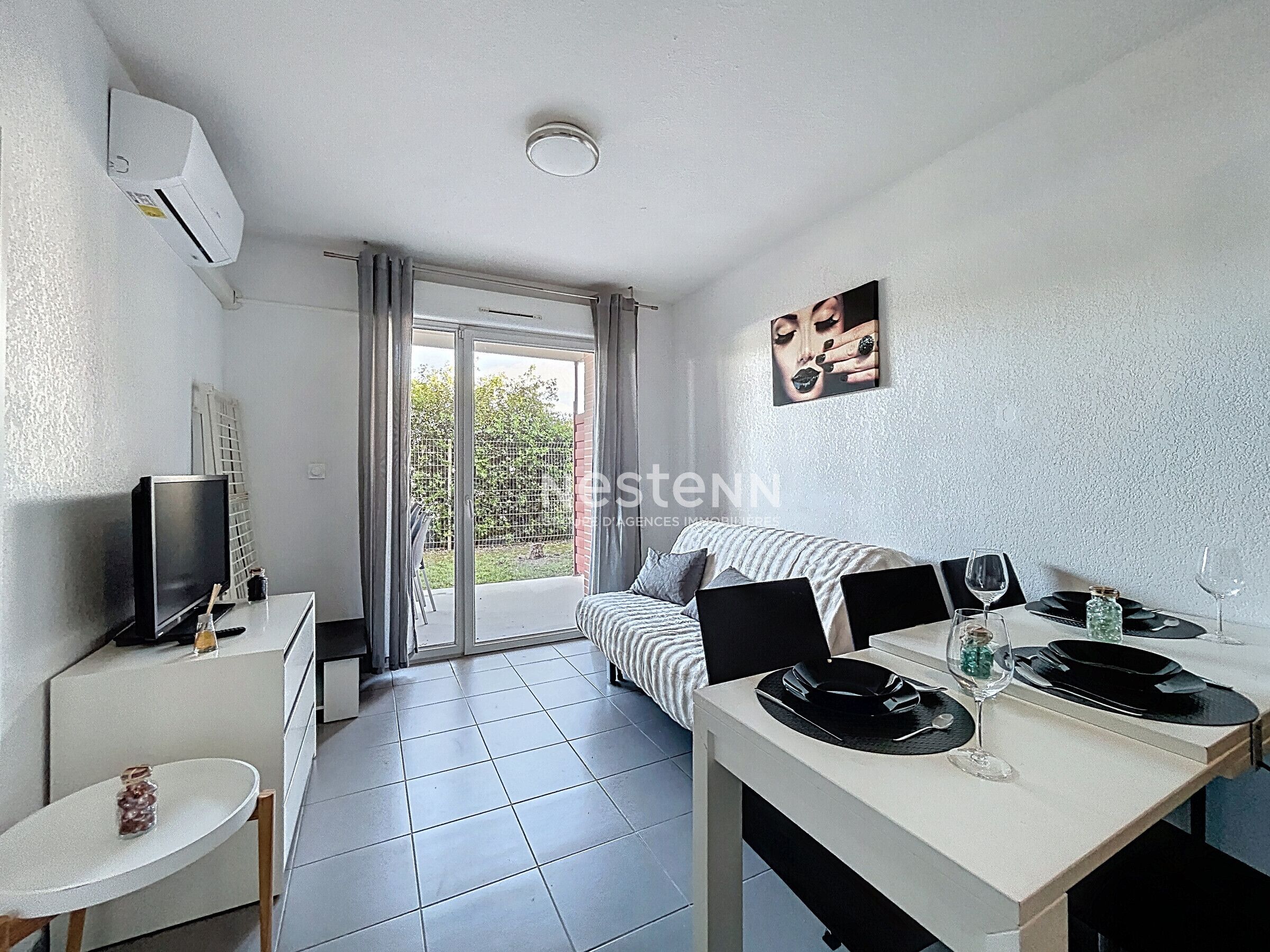 Een moderne woonkamer met een bank, een televisie, een eettafel gedekt voor twee personen en een glazen schuifdeur naar een balkon.